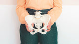 A person wearing a peach-colored blouse and green pants holds a white anatomical model of the pelvis at waist level, symbolizing pelvic health.