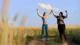 Two women with diverse body types stand outdoors in running gear, smiling and laughing. They hold hands raised in the air, celebrating a shared achievement.