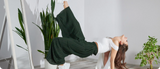A woman wearing a white cropped top and loose green pleated pants performs a dynamic, gravity-defying pose in a minimalist room surrounded by indoor plants. Her confident movement reflects freedom and control, symbolizing the empowerment and active lifestyle supported by Uresta.