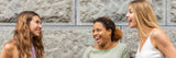 Three women laughing together in front of a stone wall, symbolizing a shared confidence, aligning with Uresta’s mission to support women with bladder leakage solutions.